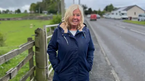 A woman with longish blonde hair in a fleece-lined long blue coat stands near a farm gate with a roadway to her left