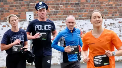 Say Cheese Studios Four runners, two men and two women, running towards the camera in Dorchester prison yard. One man and woman are dressed as police officers, the other woman is dressed in an orange prison uniform. The other man is dressed in shorts and a long-sleeve running top.