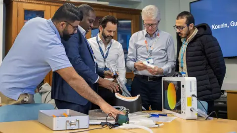 A group of men standing in a conference room, standing over a table looking at a white hospital contraption and other items. The men are dressed mainly in suits wearing lanyards.