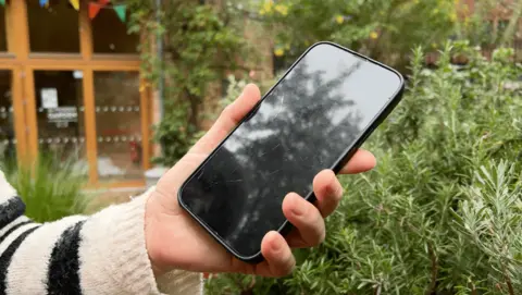 A woman wearing a black and cream jumper uses a mobile phone in a community garden. 