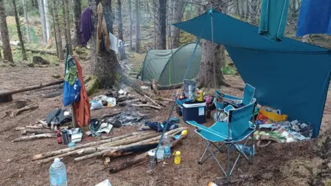 Highland Council A campsite in a forest. There is a tent in the background and a deckchair under a canopy. Food and drink containers are scattered across the ground and there are long logs collected for a fire. The embers of a campfire can also be seen. 