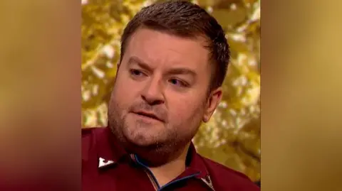 Alex Brooker in front of a gold background