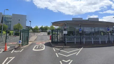 The entrance to the Benton Park View building. There is a green fence, red and white arm barrier on the road and a glass fronted building with a security guard sitting inside. A grey building can be seen in the background.