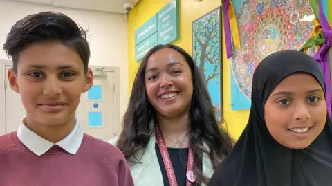 A young boy wearing a light maroon sweatshirt standing in front of a woman with long, brown hair and wearing a red lanyard who is also behind a young girl wearing a black headscarf (khimar).  
NOTE: Please pixilate school name on Rayyan's sweatshirt. 