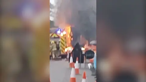 A grainy piece of footage showing a fire engine behind a burning lorry, with cones in the foreground