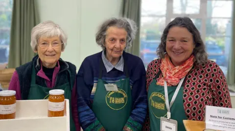 Dorothy, Jean and Christa are standing side-by-side, wearing green aprons with "Cambridgeshire county markets" written on them. There are two jars of marmalade on stands in front of them.