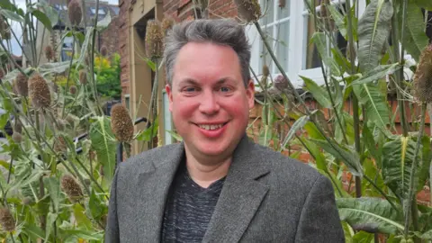 Nick Smith A man with short grey and white hair wearing a grey suit and T shirt smiles at the camera. Behind him are tall thistle plants.