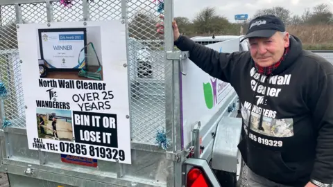 Lara King/BBC Frank Sparkes wearing a black top bearing his nickname - the North Wall Cleaner - standing at the side of a silver-coloured trailer.