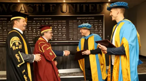 Northumbria University Eddie Howe and Dan Burn with staff from Northumbria University in the Newcastle United media room. The pair are wearing blue and gold robes. University chancellor George Clark is in a black robe and another man is in red, to his left. Howe is shaking hands with the man in red and smiling. They are standing in front of a black and white advertising board with various Newcastle sponsors and a long black desk.