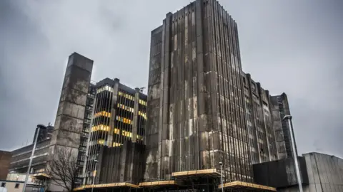 A large, multi-storey, dilapidated looking hospital building with brown and grey cladding stained with grime. 