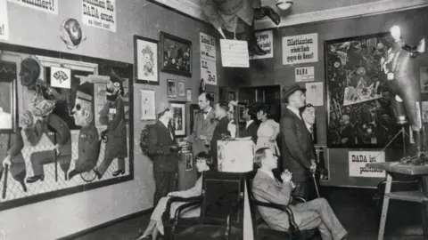 A group of people sit in an art gallery. The paintings and sculptures are all Dada. 