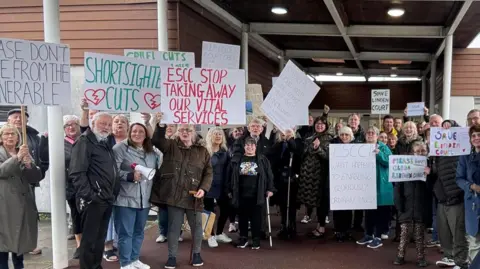 Dozens of people stood outside a large brown building while a handful hold placards with messages to stop adult social care cuts