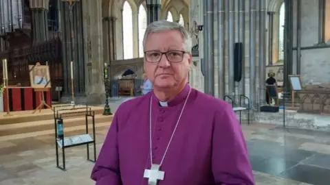 The Bishop of Salisbury stands in Salisbury Cathedral in purple clergy garments with a large cross round his neck.