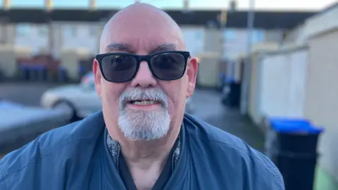 David McNicholl is wearing sunglasses. He has a goatee. he's outside. Bins are in the background. 