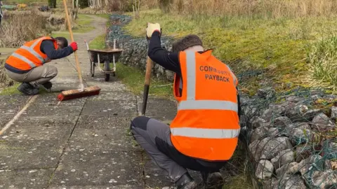 Two men in orange high visibility vests that has community payback written on the back. They are both crouched down on the floor clearing the cracks in the pavement.