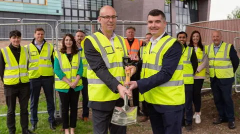 A group of people wearing yellow high visibility jackets are stood, posing for a photograph. Two men at the front of the group are holding a shovel. In the background is fencing and a large, modern-looking building.