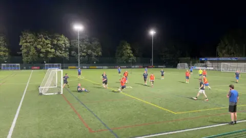 Men playing a 6-a-side football match with a black shirts team and a team wearing orange bibs. They are playing on an astro turf pitch, in the dark with the flood lights on.