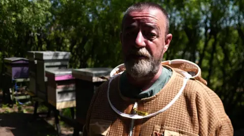 Michelle Lyons/BBC A man in a beekeeper outfit looks at the camera. There are beehives in the background.