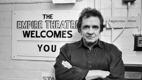 Getty Images Johnny Cash poses by standing against a wall with the sign "The Empire Theatre welcomes you" behind him