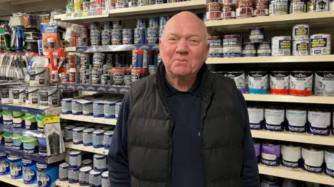 Bald man in black body warmer stood infront of shelves of paint