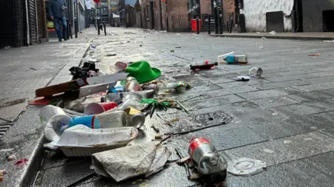 A close up of a road with a pile of litter next to the curb. The litter includes plastic bottles and cups, food boxes and a green bowlers hat