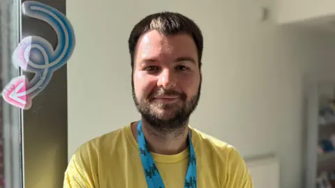 Pierce is a young man with short, brown hair and facial hair. He is smiling at the camera with his mouth closed. He is wearing a yellow t-shirt and a blue lanyard with Eastside Partnership logo on it. Behind him is a blank wall.