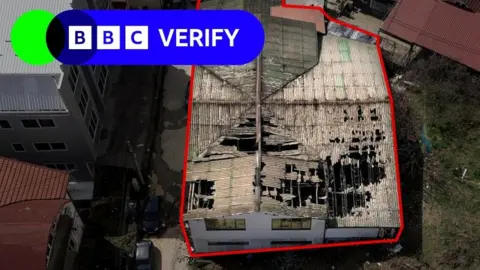 An aerial view of the damage to the Pulse nightclub in North Macedonia. It is a rectangular building resembling a former warehouse, with whitewashed walls. The roof has been badly bunt and has several large holes that reveal the skeleton of the building. The building is outlined with a thick red line to distinguish it from nearby buildings. The BBC Verify logo is over the top.