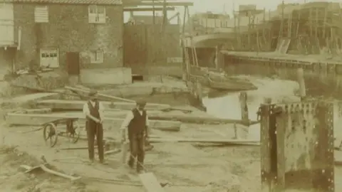 Robb Robinson A sepia image showing two men wearing Edwardian clothes walking through a shipyard on the banks of a river, with a house and numerous wooden structures in the background. A ship is being built on the far bank, surrounded by scaffolding.
