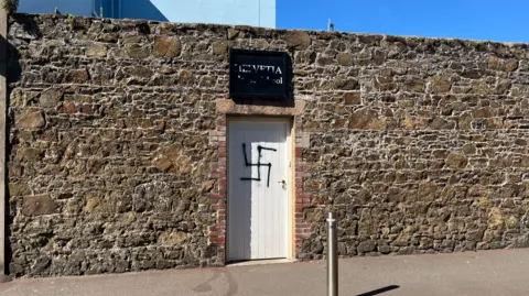 A white school door entrance on the side of the road with a black graffitied swastika on it. 