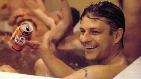 Capital Films A group of men sit swig beer from cans in a large communal bath