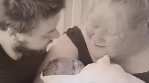 A black and white image of Lee and Kristina Pepper holding their son Theo who is wrapped in a swaddle