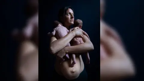 Mother Eleanor Church holding her newborn twins into her chest, showing her stomach. The twins have their backs to the camera and Ms Church is looking to the side.