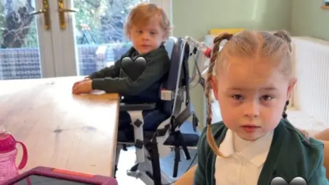 Harry and Harley are sat at the kitchen table wearing their green school uniform. They have blonde fair-coloured hair. 