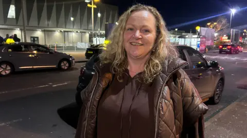 Charlotte Mawhinney is standing outside an airport. Cars are driving past. Her blond hair is curly. She's wearing a brown top and coat.