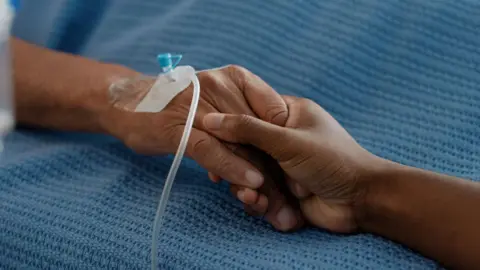 Getty Images Two people hold hands across a hospital bed. 