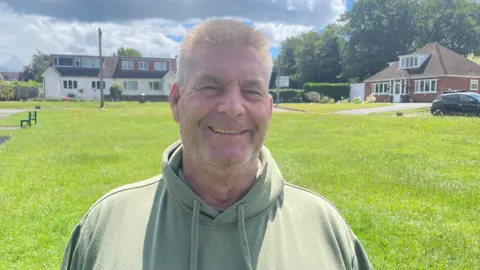 A man with short grey hair, smiling, wearing a sage green hooded top. He stands on a village green with mown grass stretching out behind him. Detached houses, trees and a village hall sign line the far edge under a cloudy blue sky.