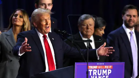 Donald Trump speaks behind a podium with his arms open - he wears a blue suit, white shirt and red tie