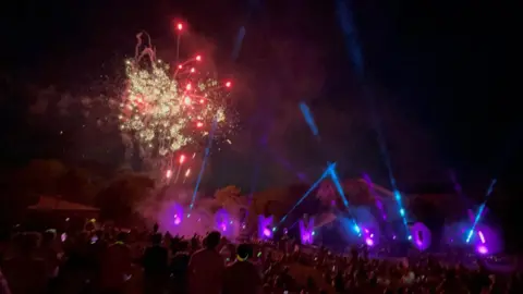 Las luces estroboscópicas de Nick Hutson se pueden ver que emanan de lugares alrededor del letrero de Oakwood. Son de color azul y morado. Se puede ver una exhibición de fuegos artificiales al fondo con colores amarillos y rojos en el cielo. Se puede ver un gran grupo de personas en el primer plano de la imagen que mira la pantalla.