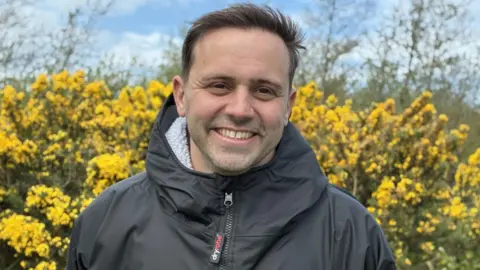 Neil smiling in a black jacket against a backdrop of yellow gorse