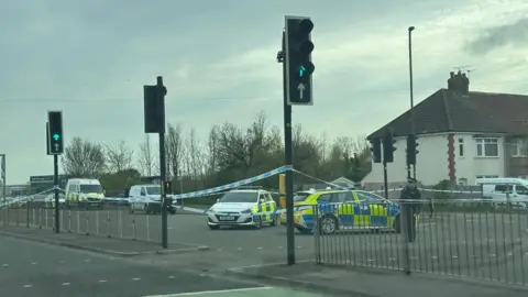 Two police cars and two vans block the road near a residential street. There is blue and white police tape across the road. 