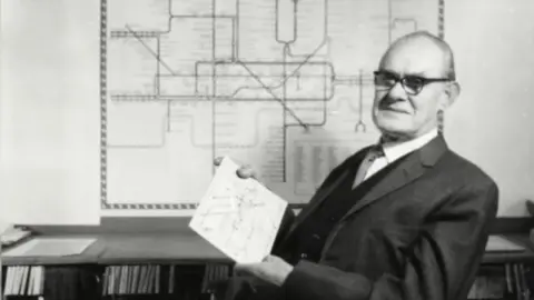 London Transport Museum A black and white image of Harry Beck wearing a suit and sitting in front of a London Underground map which is on the wall behind him