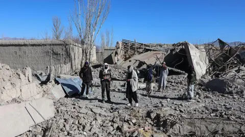 AFP via Getty Images Men walk around the area where a house was destroyed in Urgun district, Pakitka province. The building's roof is completely collapsed and rubble from the building covers the ground.