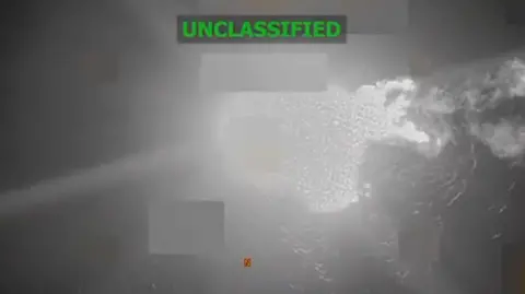 US Department of Defense What appears to be a black and white image of the sea as seen from above. A few areas are blurred, including the section where the boat is. A large white area which appears to be the flames of an explosion are in the centre of the image. The word "unclassified" is captioned on the image. 
