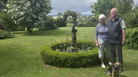 David and Liz stood on a large mown lawn with bushes and trees in the background - arranged in a landscaped fashion. At their feet is a small border terrier.