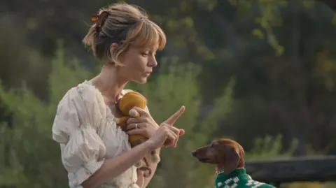 Netflix Carey Mulligan in Beef 2, wearing a white patterned dress and holding a finger up to to a dog, who is wearing a green jumper 