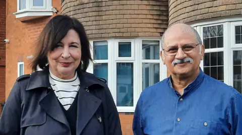 A woman and a man standing on a driveway in front two residential properties. The woman is in a navy trench coat and the man is in a denim shirt. They are both smiling into the camera.