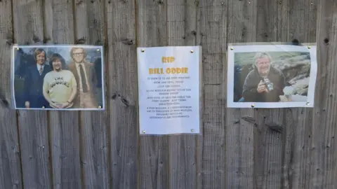 Tom Johnston Three laminated sheets on a wooden fence. On the left, a picture of the Goodies - three men in seventies clothing.. two in suits flanking Bill Oddie in a Goodies T-shirt at the front. In the middle, a sign headed "RIP Bill Oddie" which mentions his link to the street and his roles in the Goodies and wildlife programmes, and a photo on the right of Oddie holding a camera during a wildlife programme.