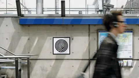 Thierry Bal A small plaque is attached to the grey concrete wall of a modern Tube station. It has a black and white image that resembles a labyrinth on it. A person walks past, his image is blurred. 