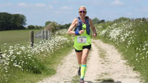 A woman, Laura Watts, in a yellow vest, black shorts and yellow high socks running along a gravel track, towards the camera. She is wearing sunglasses, has tied back blonde hair and is wearing blue straps across her shoulders. Either side of the track is green hedgerow with white flowers. To the left of the image is a green field with large green trees in the background. The sky is blue with a few clouds.