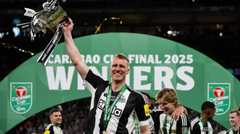 Getty Images Dan Burn after winning the Carabao Cup. He's wearing a black and white Newcastle United top, with a green medal around his neck. He's lifting the silver trophy in one hand which has black and white ribbons and standing in front of a green arch which reads 'Carabao Cup Final 2025 Winners'. He's smiling into the camera.